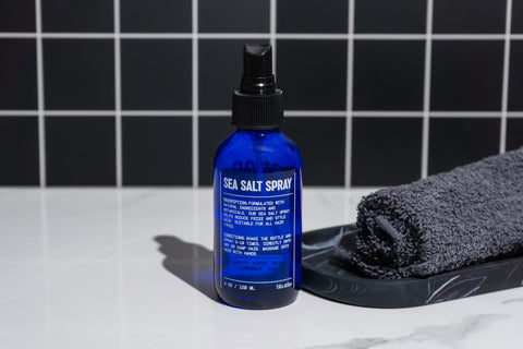 A blue bottle of Blu Atlas Sea Salt Spray, famed for its texture-enhancing abilities and natural components, rests on a marble surface next to a rolled black towel. The black and white tile walls in the background create an elegant contrast.