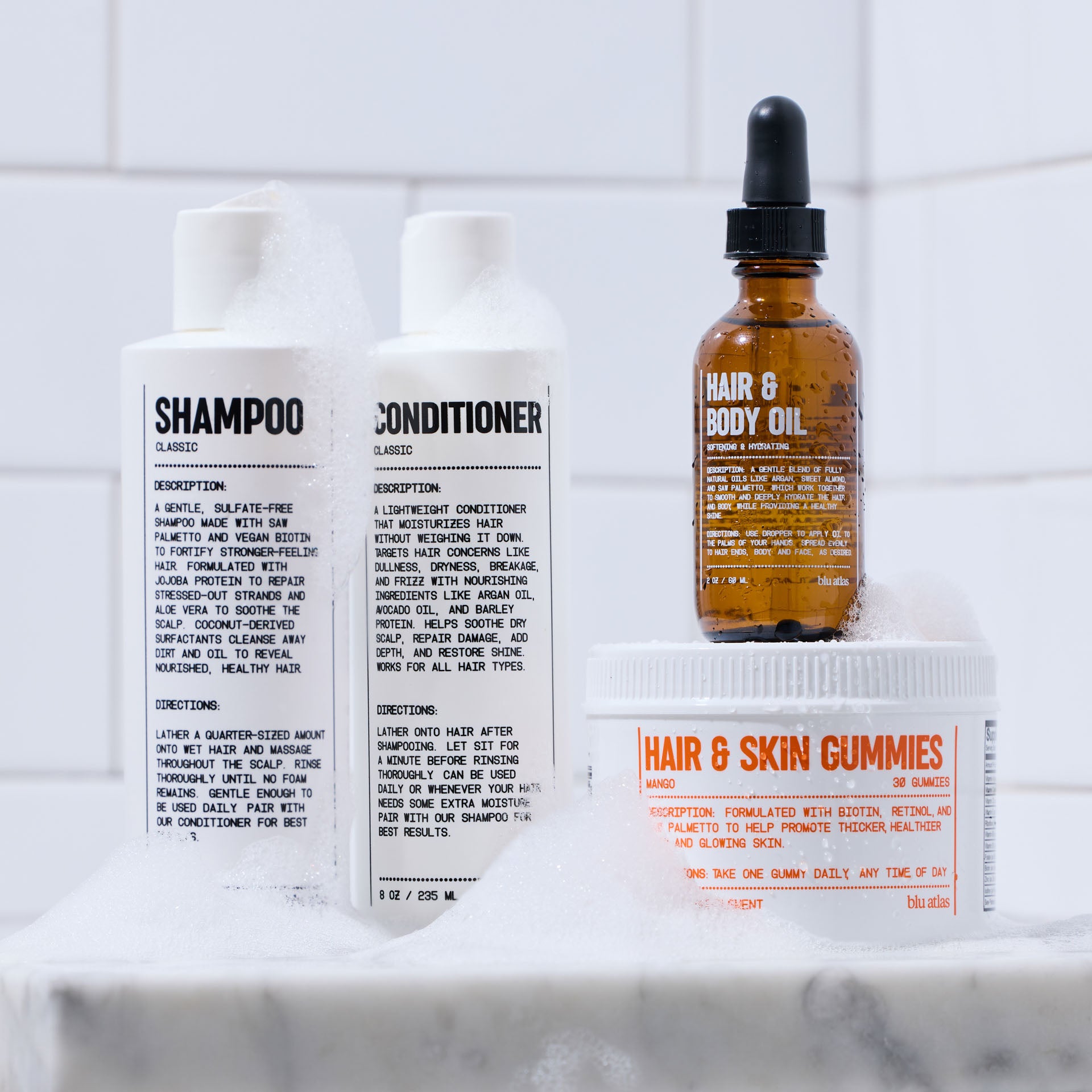 On a wet surface, the Ultimate Hair Care Set by Blu Atlas features four essentials: shampoo, conditioner, hair & body oil, and hair & skin gummies. Bubbles elegantly shimmer around the products, highlighting their clean ingredients. This set makes an ideal gift for anyone who values natural beauty and nourished hair and scalp.