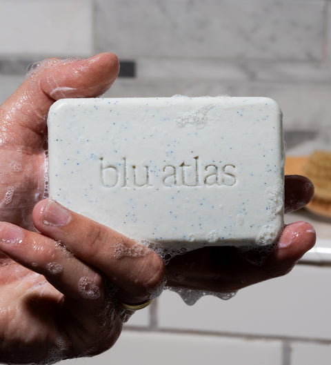 A close-up of a person's hand displays the Blu Atlas Natural Exfoliating Bar Soap, engraved with the brand's name. The soap, enriched with jojoba beads, is covered in lathered bubbles.