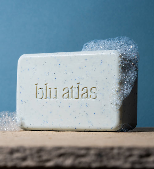 A bar of Blu Atlas Natural Exfoliating Bar Soap, enriched with nourishing shea butter and gentle jojoba beads, with blue specks and rich foam, rests on a stone surface against a blue background.
