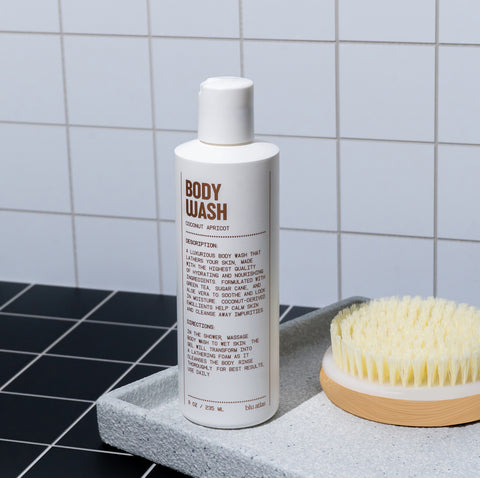 The Blu Atlas Hydrating Body Wash, labeled Coconut Apricot, rests on a gray tray with a brush against a tiled bathroom wall. Enriched with coconut-derived emollients, it hydrates skin, leaving it soft and pampered.