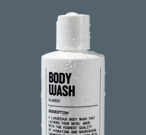 A white bottle labeled Hydrating Body Wash by Blu Atlas, adorned with water droplets against a gray background, describes a luxurious formula enriched with coconut-derived emollients for nourishing hydration.