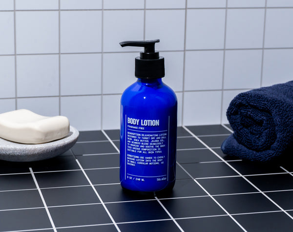 A blue bottle of Blu Atlas Natural Body Lotion with shea butter and a pump sits on a black tiled counter next to a bar of soap and a folded navy towel.