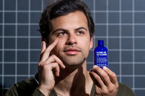 A person holds a blue bottle labeled Blu Atlas Volcanic Ash Gel Cleanser, an exfoliating face wash for all skin types, and applies it while standing before a grid-patterned background.