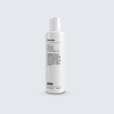 White cylindrical bottle from Blu Atlas, labeled as Natural Shampoo for All Hair Types. The packaging highlights vegan biotin and jojoba protein and emphasizes that it is vegan and cruelty-free, complete with a detailed ingredient list.