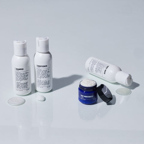 Four toiletry bottles, including shampoo, conditioner, face wash, and a blue jar of face moisturizer, are placed on a light surface with some product spilled beside each container.