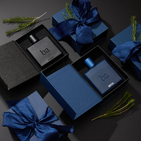 Two cologne bottles, one black and one blue, are displayed in open gift boxes with blue ribbon, surrounded by sprigs of greenery on a dark surface.