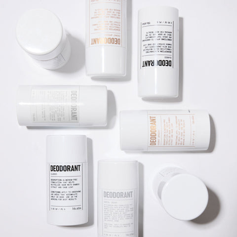 Several white cylindrical deodorant containers are arranged on a white surface, with minimalist black and orange text labels visible on some of them.