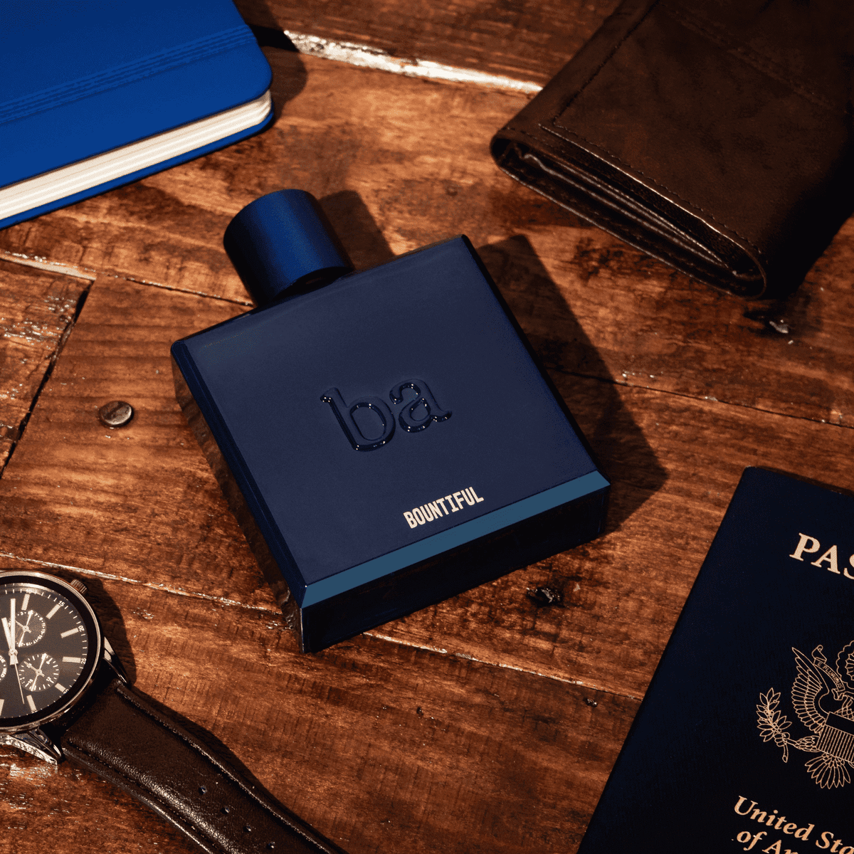 Two cologne bottles, one black and one blue, are displayed in open gift boxes with blue ribbon, surrounded by sprigs of greenery on a dark surface.
