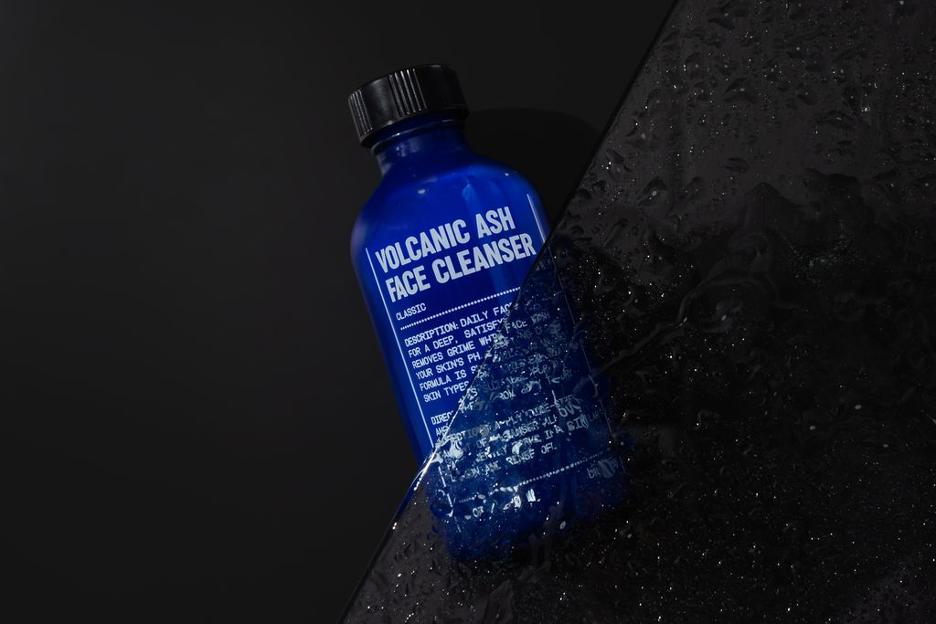 Three blue skincare containers labeled: Volcanic Ash Face Cleanser, Exfoliating Scrub, and Moisturizing Cream, placed on a black surface.