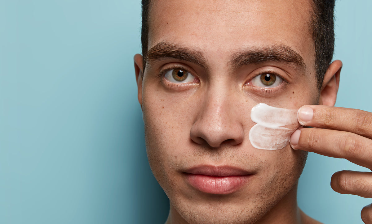 A person applies a small amount of cream to their cheek against a blue background.