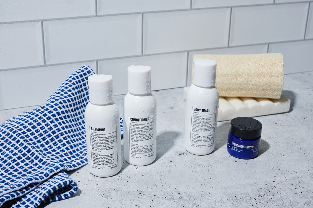 Three small bottles labeled shampoo, conditioner, and body wash from the Blu Atlas Essentials Travel Set are arranged next to a blue cloth, a face moisturizer jar, and a bath sponge on a gray countertop with a white tile background.