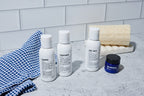 Three small bottles labeled shampoo, conditioner, and body wash from the Blu Atlas Essentials Travel Set are arranged next to a blue cloth, a face moisturizer jar, and a bath sponge on a gray countertop with a white tile background.