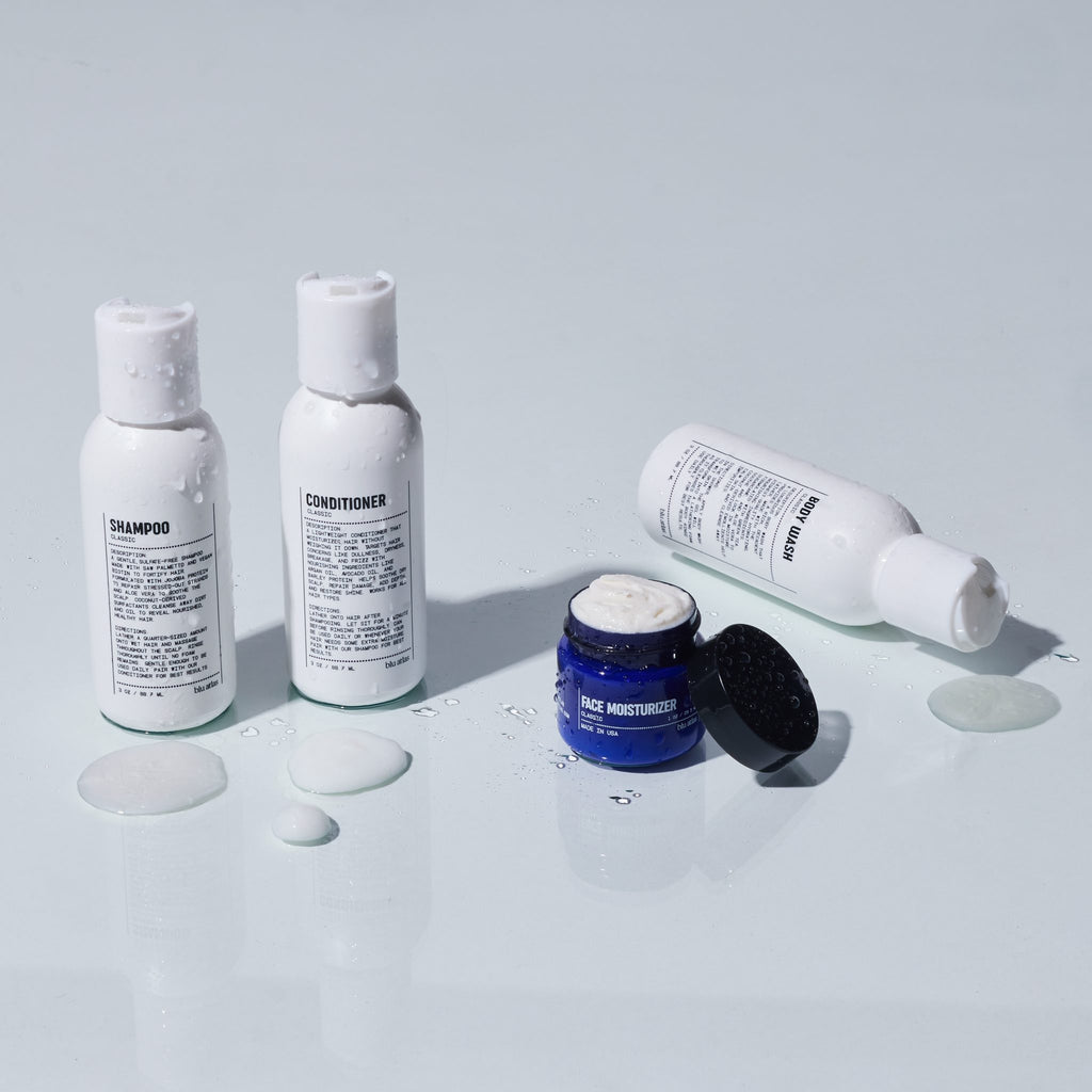 Three bottles labeled shampoo, conditioner, and moisturizer—accompanied by a blue face moisturizer jar—are displayed with spilled product on a white surface, showcasing the convenience of the Blu Atlas Essentials Travel Set.