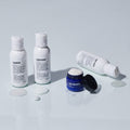 Three bottles labeled shampoo, conditioner, and moisturizer—accompanied by a blue face moisturizer jar—are displayed with spilled product on a white surface, showcasing the convenience of the Blu Atlas Essentials Travel Set.