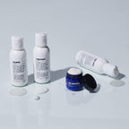 Three bottles labeled shampoo, conditioner, and moisturizer—accompanied by a blue face moisturizer jar—are displayed with spilled product on a white surface, showcasing the convenience of the Blu Atlas Essentials Travel Set.