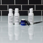 Four products from the Blu Atlas Essentials Travel Set—a trio of white bottles labeled Shampoo, Conditioner, and Body Wash—are displayed behind an open jar marked Face Moisturizer on a reflective surface with a black tiled background.