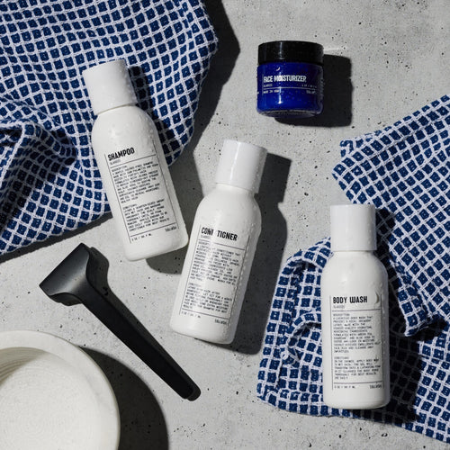 Three white bottles labeled shampoo, conditioner, and body wash from the Blu Atlas Essentials Travel Set, a blue face moisturizer jar, a black razor, and blue checkered towels are arranged on a gray surface.