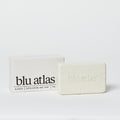 A rectangular Blu Atlas Exfoliating Bar Soap, embossed with the brand and placed before a matching white box labeled "Blu Atlas Classic Exfoliating Bar Soap 7oz." Formulated with shea butter for nourishment and gentle exfoliation.