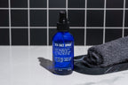 A blue bottle of Blu Atlas Sea Salt Spray, famed for its texture-enhancing abilities and natural components, rests on a marble surface next to a rolled black towel. The black and white tile walls in the background create an elegant contrast.