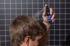 An individual uses Blu Atlas Sea Salt Spray, which contains natural ingredients, to enhance their hair's texture while standing in front of a sleek black tiled wall.