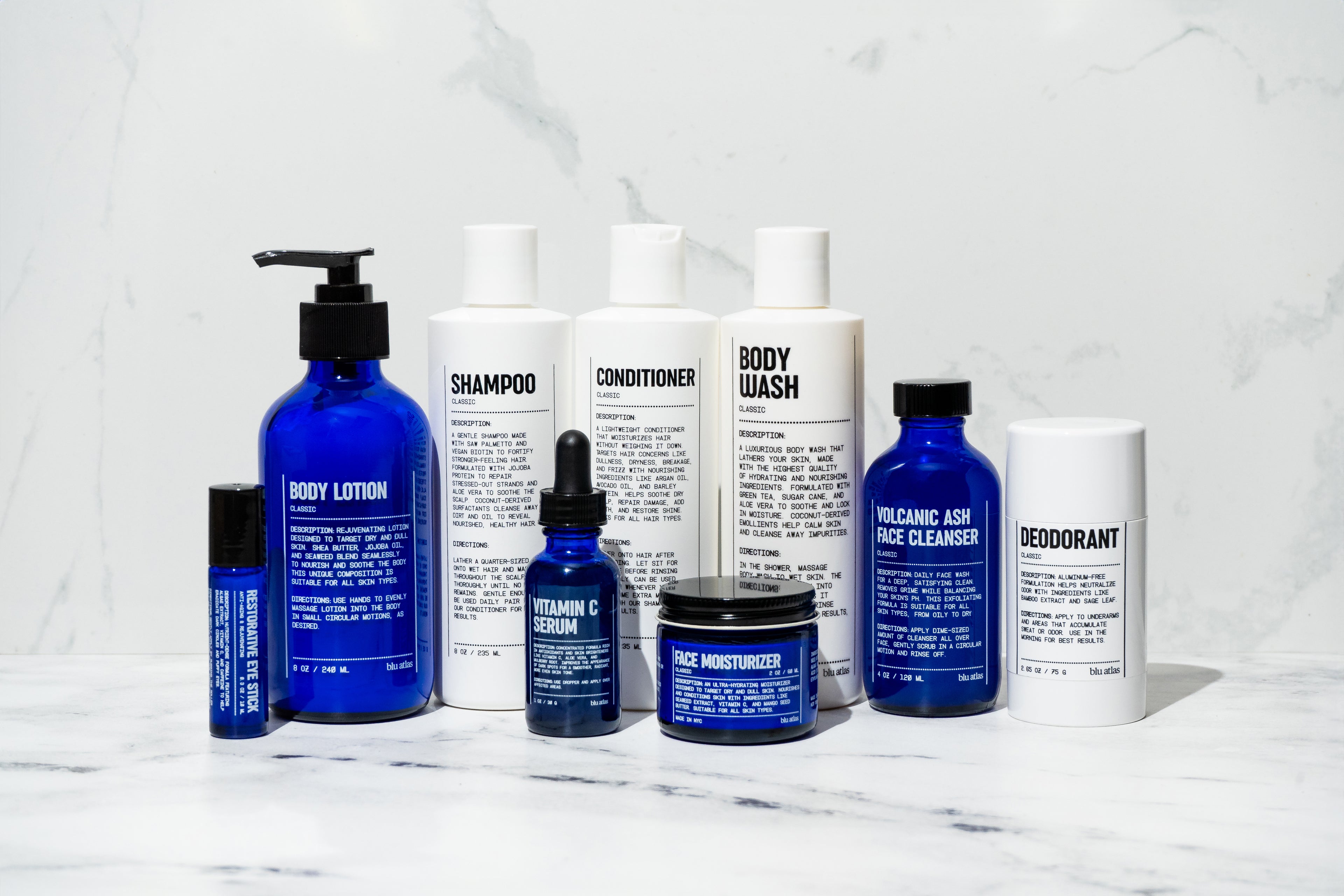 Assorted blue and white skincare products including lotions, shampoo, conditioner, body wash, face cleanser, serum, and deodorant on a marble surface.