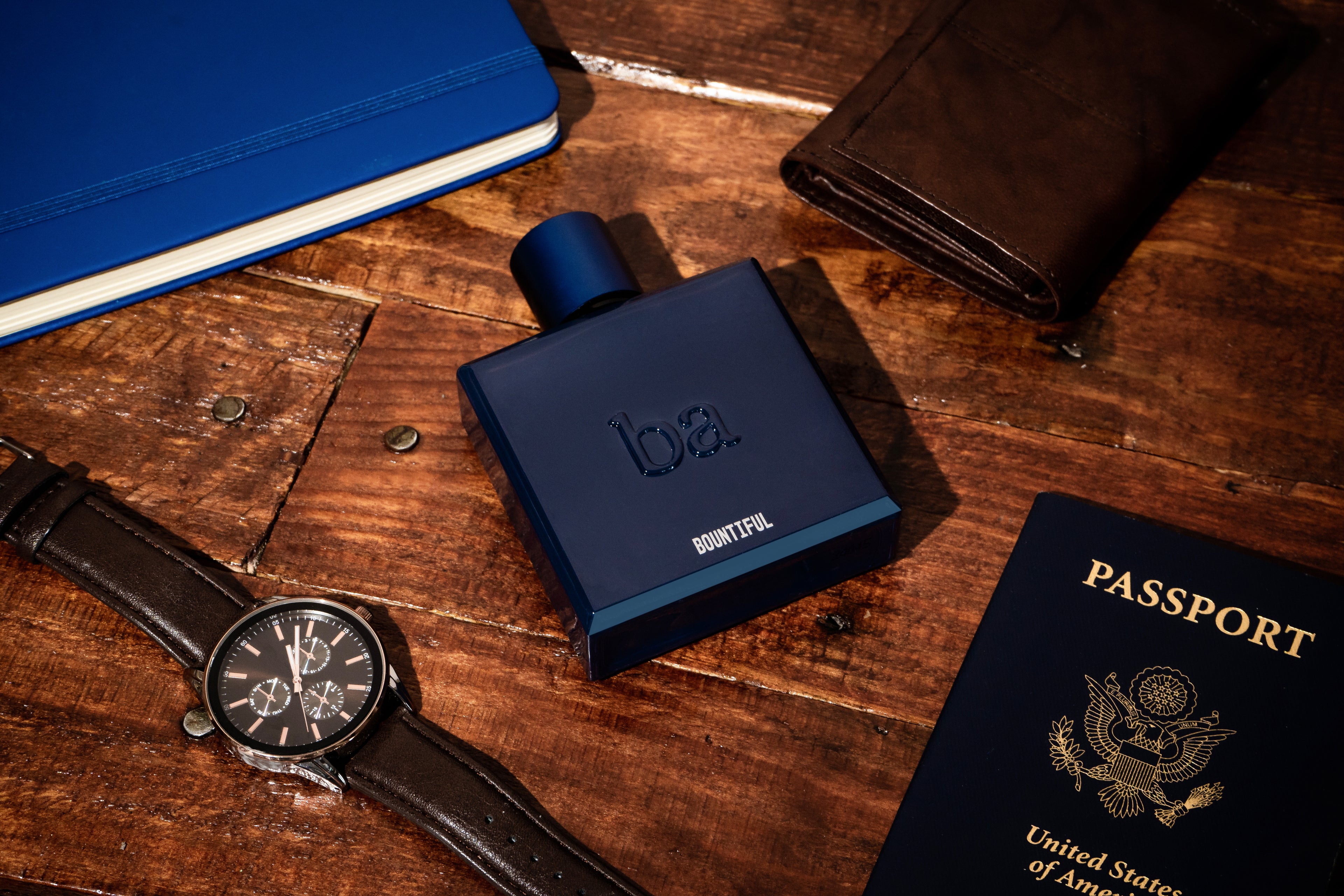 Flat lay of a blue notebook, brown leather wallet, blue Bountiful Eau de Parfum by Blu Atlas, wristwatch, and a U.S. passport on a wooden surface—ideal elements for crafting the perfect holiday gift ensemble.