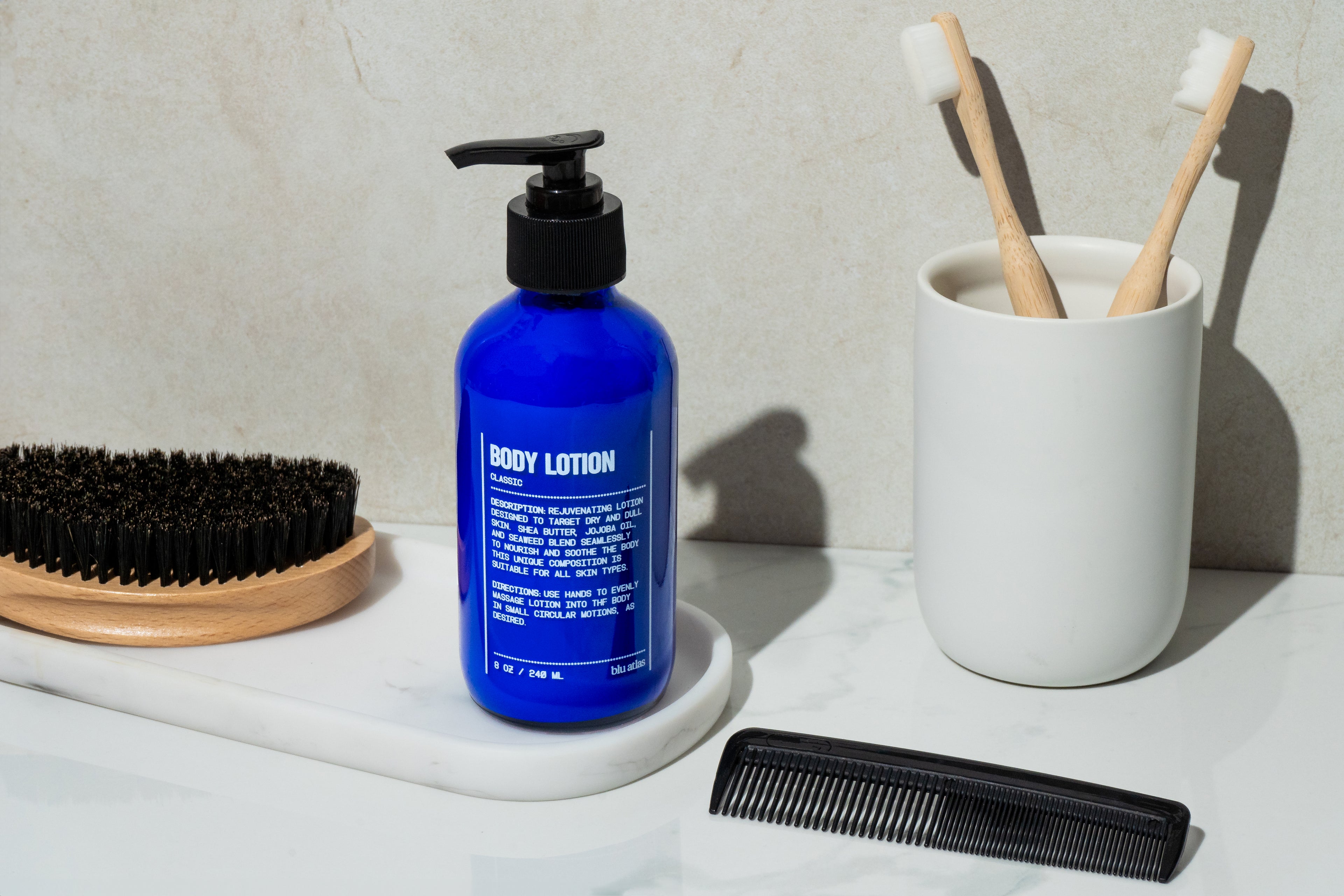 A bottle of Blu Atlas Body Essentials stands on a white surface beside a black brush, black comb, and a cup with two bamboo toothbrushes, creating an inviting scene centered on skin hydration essentials.