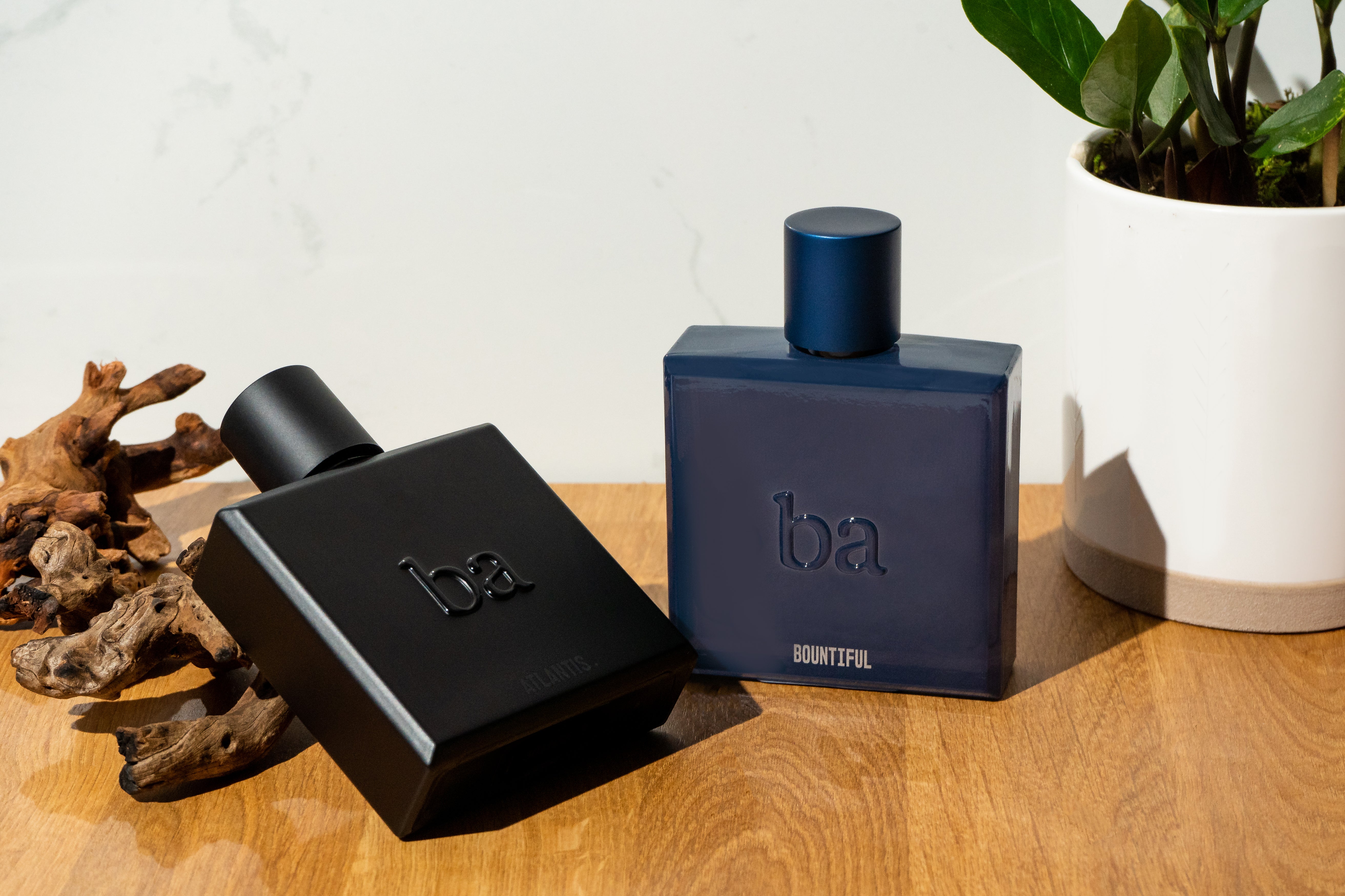 Two bottles labeled Joonie Aftershave 6 from Blu Atlas rest on a wooden surface alongside a potted plant and driftwood, highlighting their naturally sourced ingredients.
