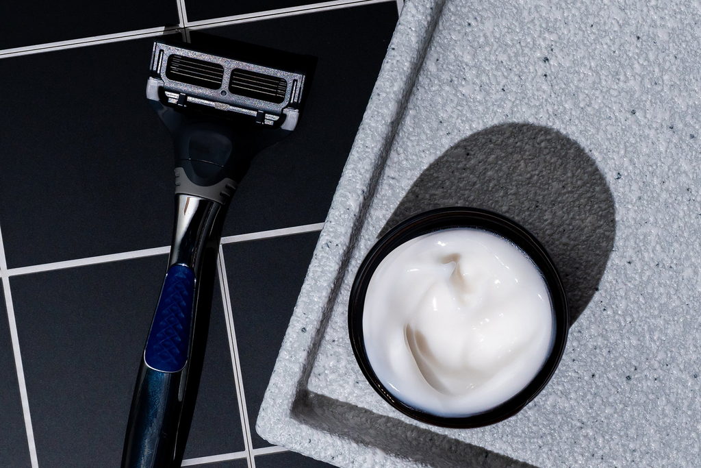 A black and silver razor rests beside an open jar of Blu Atlas Shave Essentials (Shave Cream + Aftershave) with its signature stearic-acid cushion, all arranged on a gray tray over a black-tiled surface.