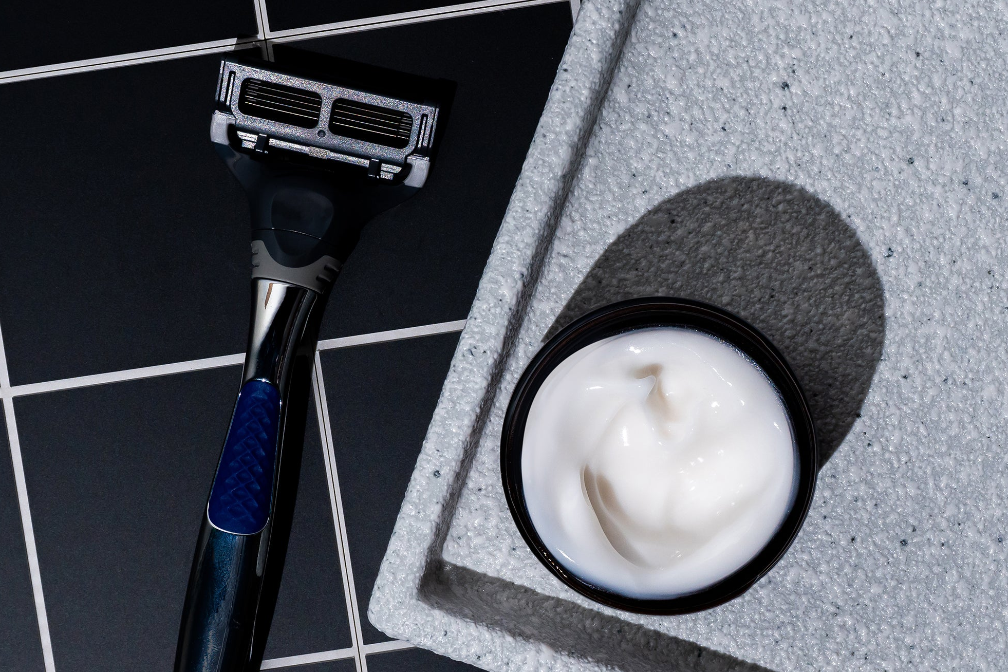 A black and silver razor rests beside an open jar of Blu Atlas Shave Essentials (Shave Cream + Aftershave) with its signature stearic-acid cushion, all arranged on a gray tray over a black-tiled surface.