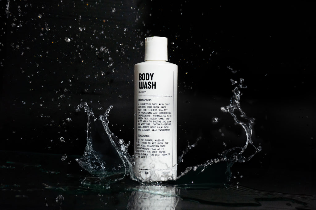A white bottle labeled Blu Atlas Body Essentials stands upright on a reflective black surface with water splashing around it, emphasizing its skin hydration benefits.