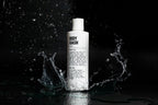 A white bottle labeled Blu Atlas Body Essentials stands upright on a reflective black surface with water splashing around it, emphasizing its skin hydration benefits.