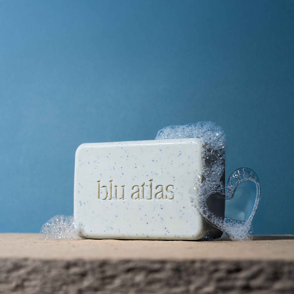 A rectangular Blu Atlas Body Care Essentials Set soap bar rests on stone with bubbles, including a heart-shaped one, highlighting natural ingredients against a blue background.