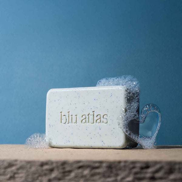A rectangular Blu Atlas Body Care Essentials Set soap bar rests on stone with bubbles, including a heart-shaped one, highlighting natural ingredients against a blue background.
