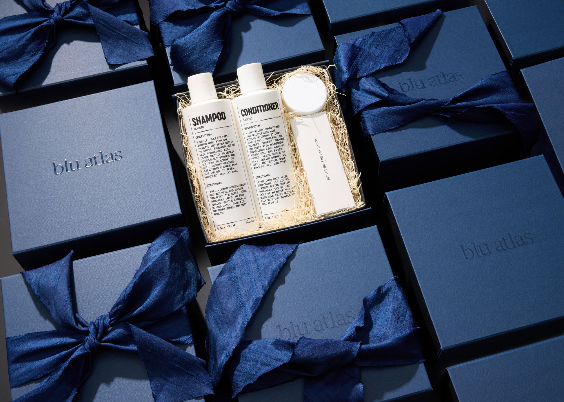 The Body Care Essentials Set by Blu Atlas includes shampoo, conditioner, deodorant, and a jar of cream. Each gift box rests on shredded paper and is among blue boxes tied with blue ribbons.