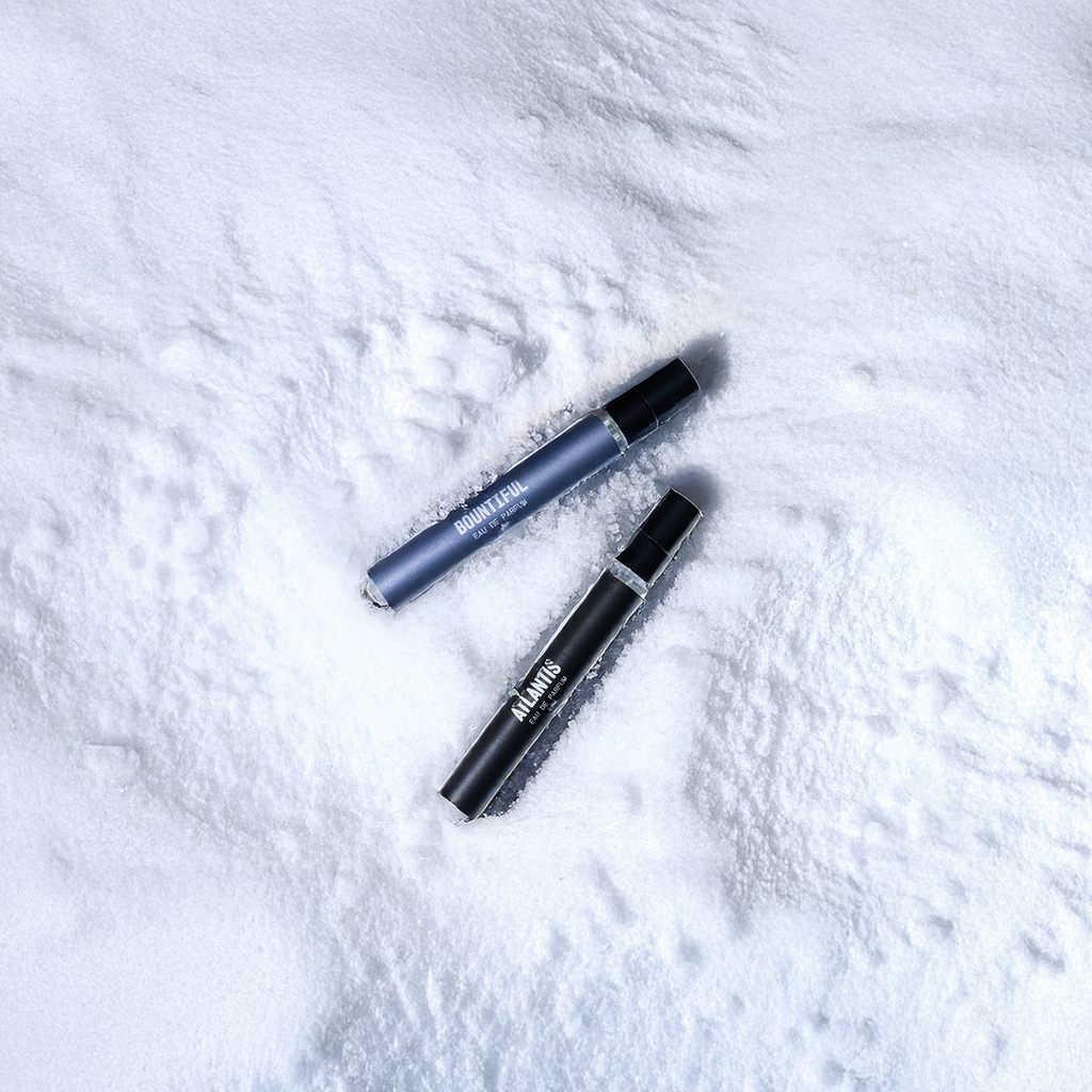 Two cylindrical cosmetic tubes from the Blu Atlas Fragrance Discovery Set, one blue and one black, rest on white snow.