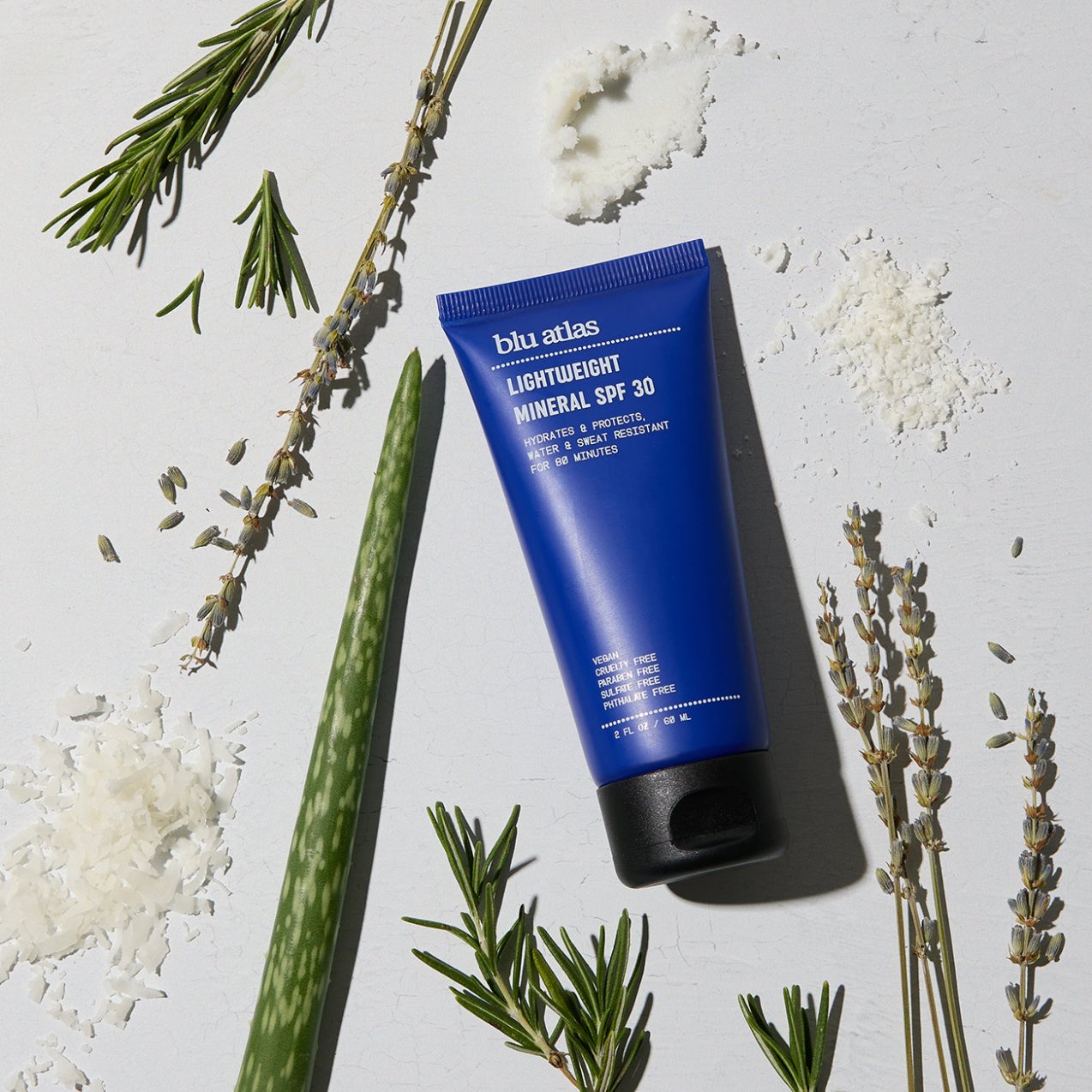 A tube of Blu Atlas Protect & Moisturize Duo rests on a light surface, surrounded by aloe, rosemary, lavender, and flakes. Perfect for natural skincare enthusiasts seeking UVA/UVB protection with water- and sweat-resistant SPF 30.