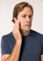 A person in a blue shirt applies Blu Atlas Lightweight Mineral SPF 30 to their cheek against a plain background.