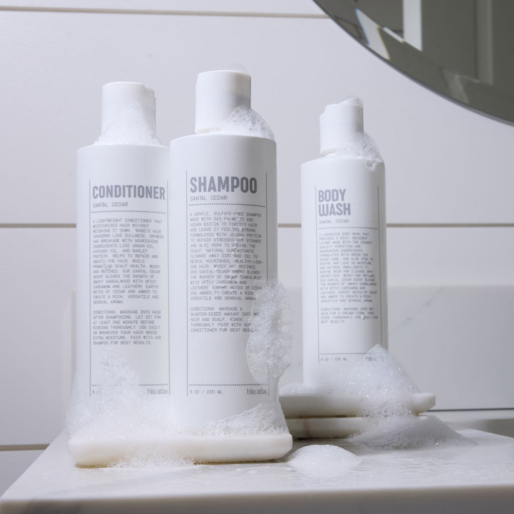 Three white bottles labeled conditioner, shampoo, and body wash with sandal cedar scent, covered in soap suds, sit on a bathroom counter next to a mirror—perfect for your daily hair care routine with Blu Atlas Hair Essentials (Shampoo + Conditioner), featuring a vegan biotin-infused formula.