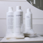 Three white bottles labeled conditioner, shampoo, and body wash with sandal cedar scent, covered in soap suds, sit on a bathroom counter next to a mirror—perfect for your daily hair care routine with Blu Atlas Hair Essentials (Shampoo + Conditioner), featuring a vegan biotin-infused formula.