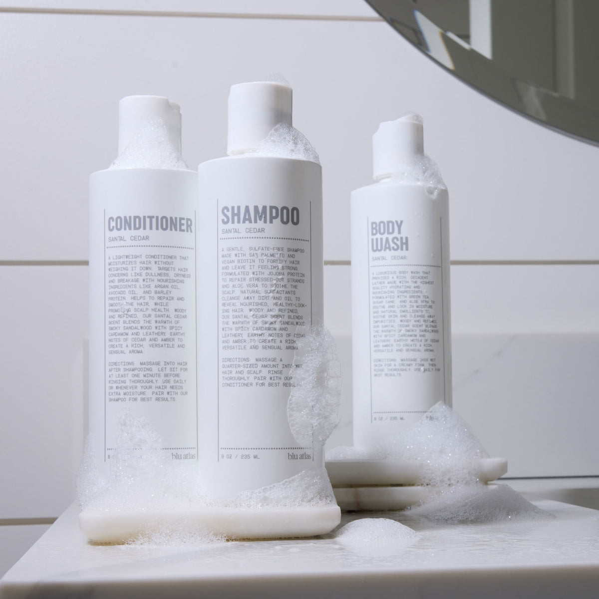 Three white bottles labeled conditioner, shampoo, and body wash with sandal cedar scent, covered in soap suds, sit on a bathroom counter next to a mirror—perfect for your daily hair care routine with Blu Atlas Hair Essentials (Shampoo + Conditioner), featuring a vegan biotin-infused formula.