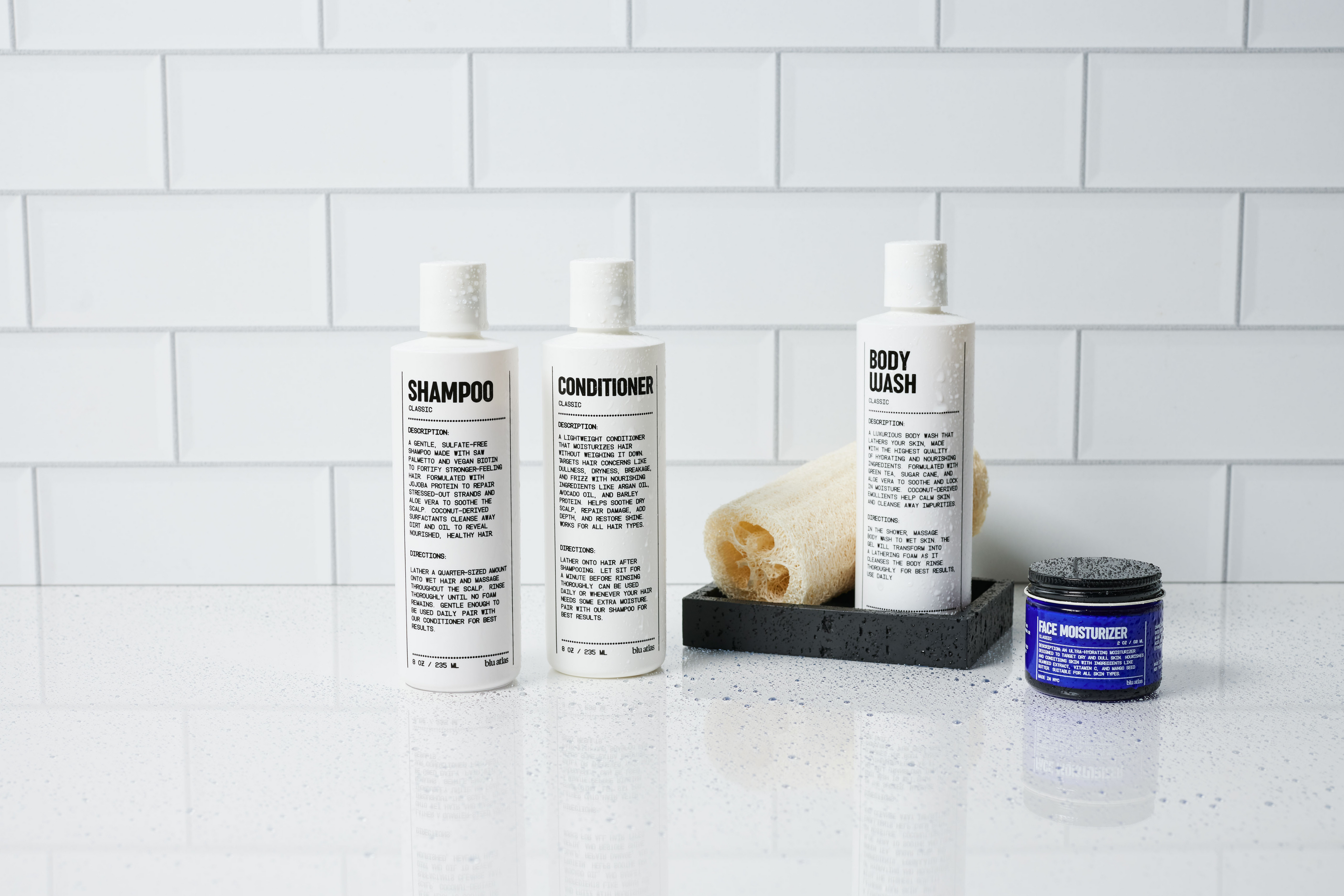 White bathroom products labeled shampoo, conditioner, and body wash are arranged on a counter with a rolled towel. A small blue jar labeled face moisturizer is nearby. White tiled background.