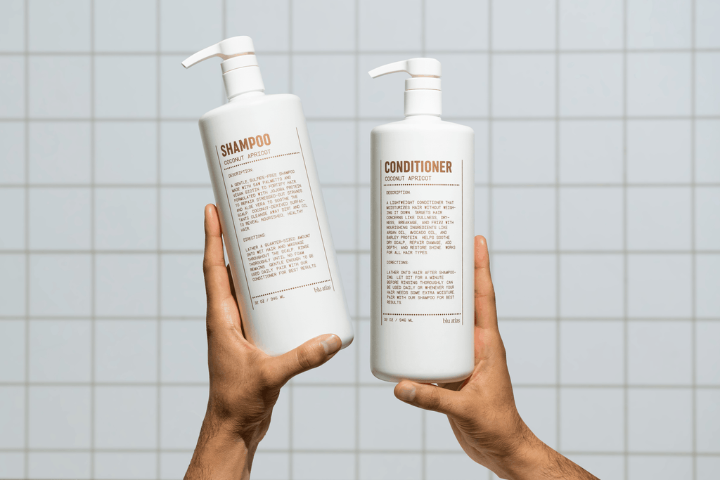 Two hands hold up white Blu Atlas pump bottles labeled Natural Shampoo - Coconut Apricot, with description text, against a tiled background.