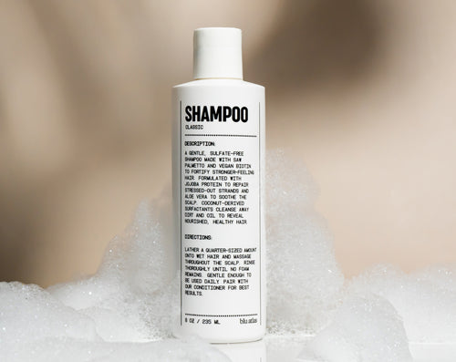 A white bottle of Blu Atlas Natural Shampoo stands upright in a pile of white soap bubbles, with the label clearly facing forward.
