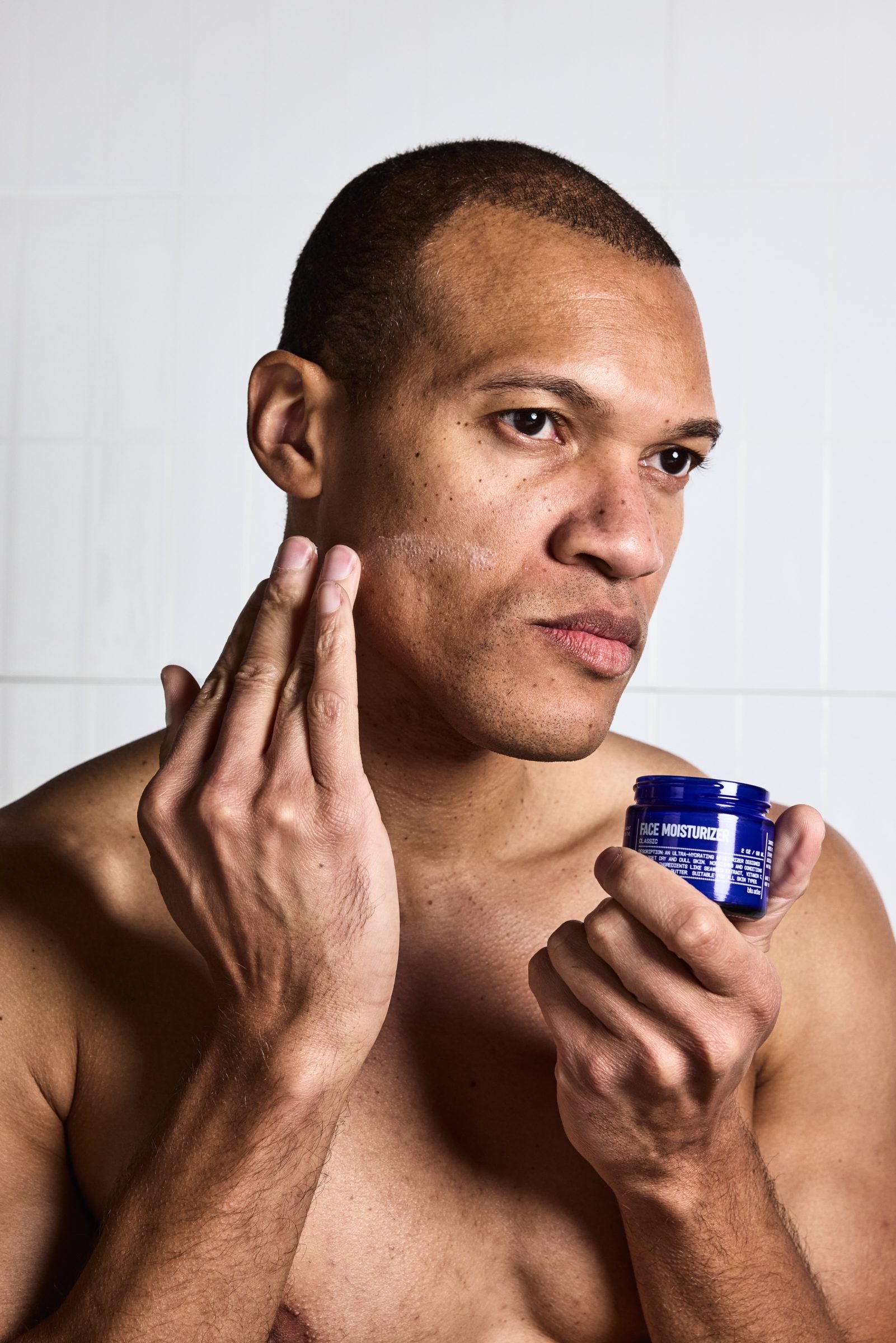 A man stands against a white tiled background, applying Blu Atlas Face Essentials (Cleanser + Moisturizer) to his cheek with one hand while holding the blue jar in the other.