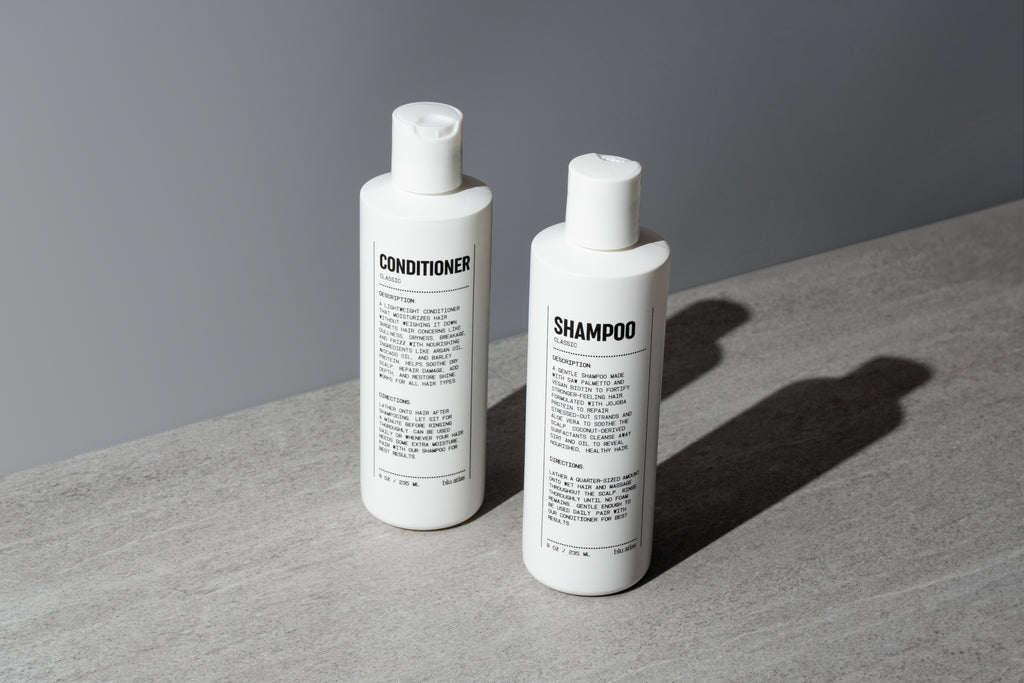 Blu Atlas Hair Essentials (Shampoo + Conditioner): Two sleek white bottles with black text rest on a gray surface, their vegan biotin formulas nourishing and strengthening your hair—the ideal daily duo for healthy strands.