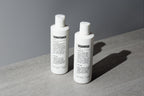 Blu Atlas Hair Essentials (Shampoo + Conditioner): Two sleek white bottles with black text rest on a gray surface, their vegan biotin formulas nourishing and strengthening your hair—the ideal daily duo for healthy strands.