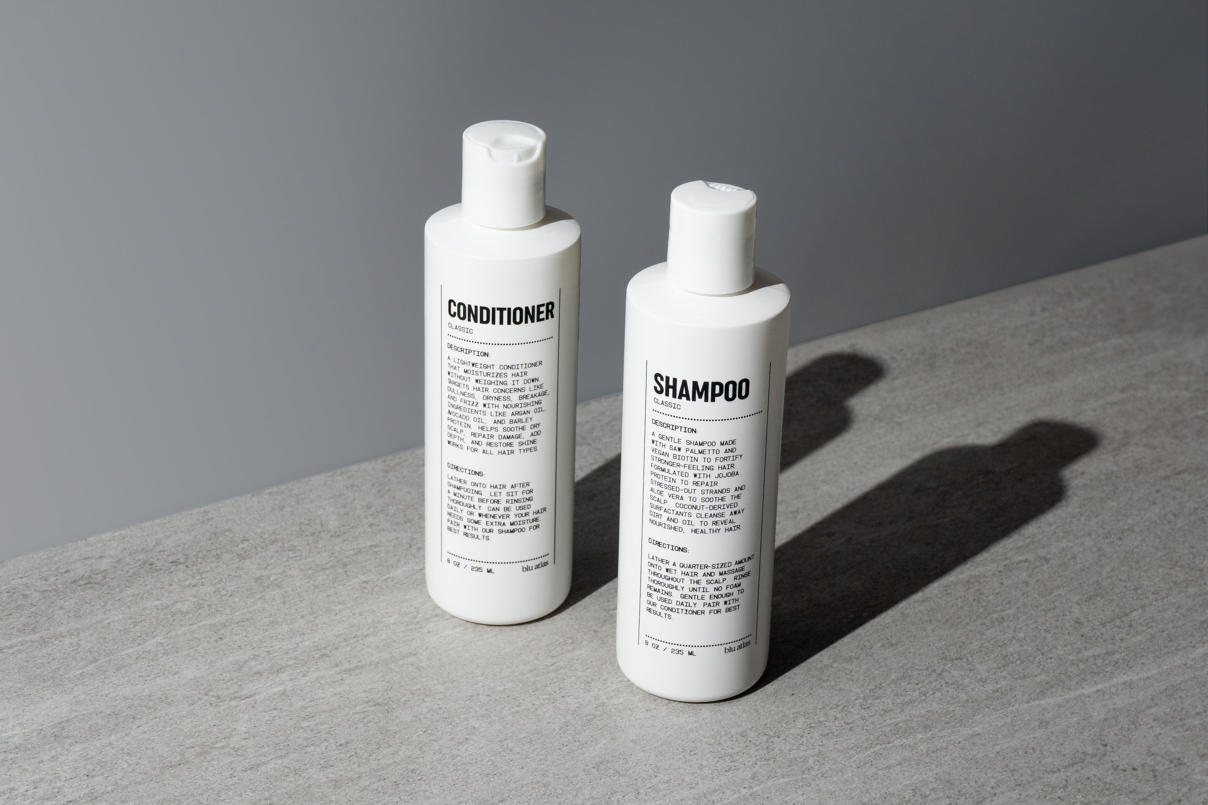 Blu Atlas Hair Essentials (Shampoo + Conditioner): Two sleek white bottles with black text rest on a gray surface, their vegan biotin formulas nourishing and strengthening your hair—the ideal daily duo for healthy strands.