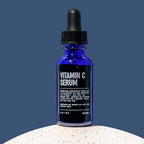 A blue glass dropper bottle of Blu Atlas Vitamin C Serum stands upright on a white textured surface against a plain blue background, showcasing its brightening ascorbic acid formula.